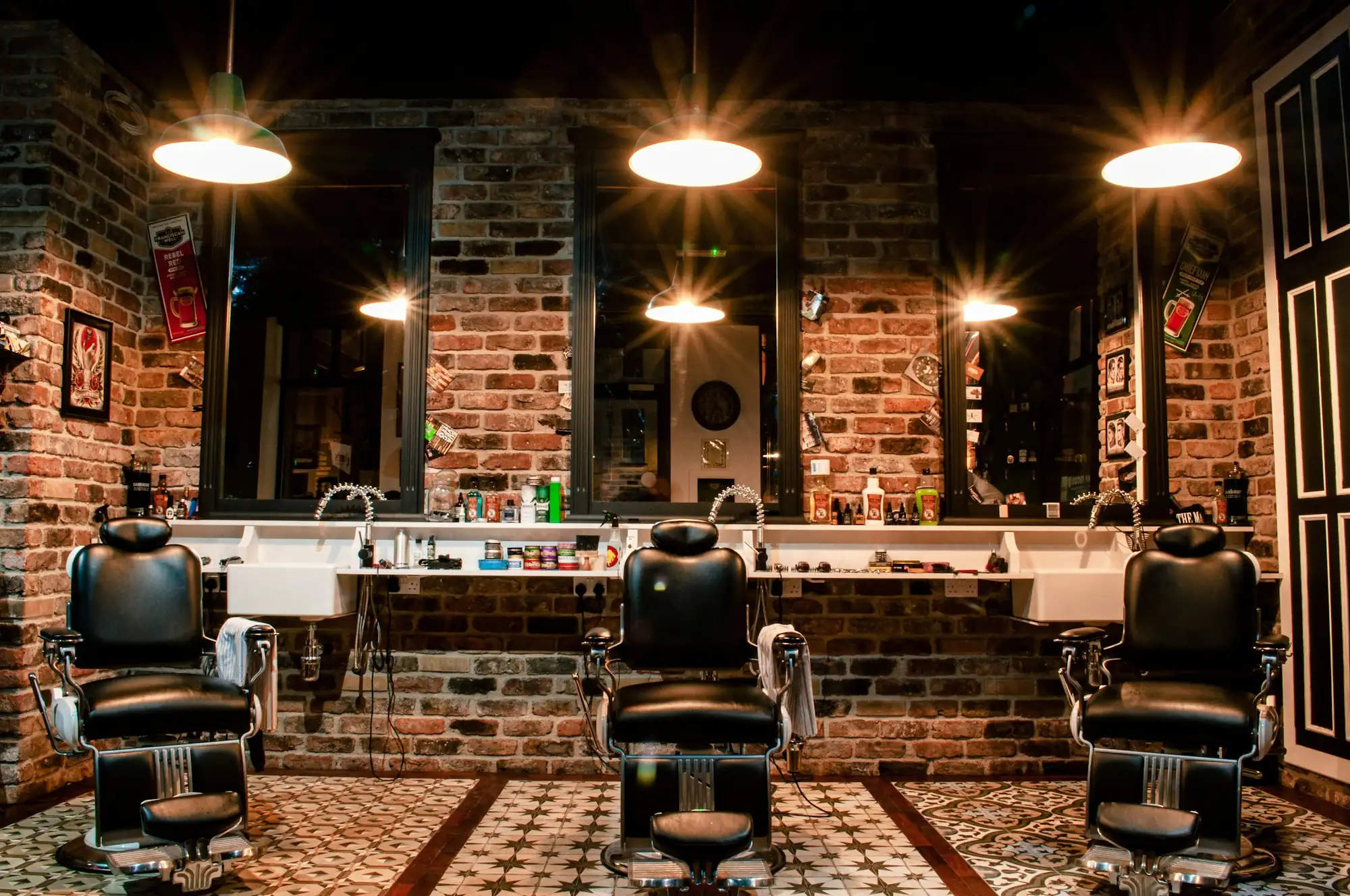 A barbershop with three leather chairs under warm pendant lights and a brick wall