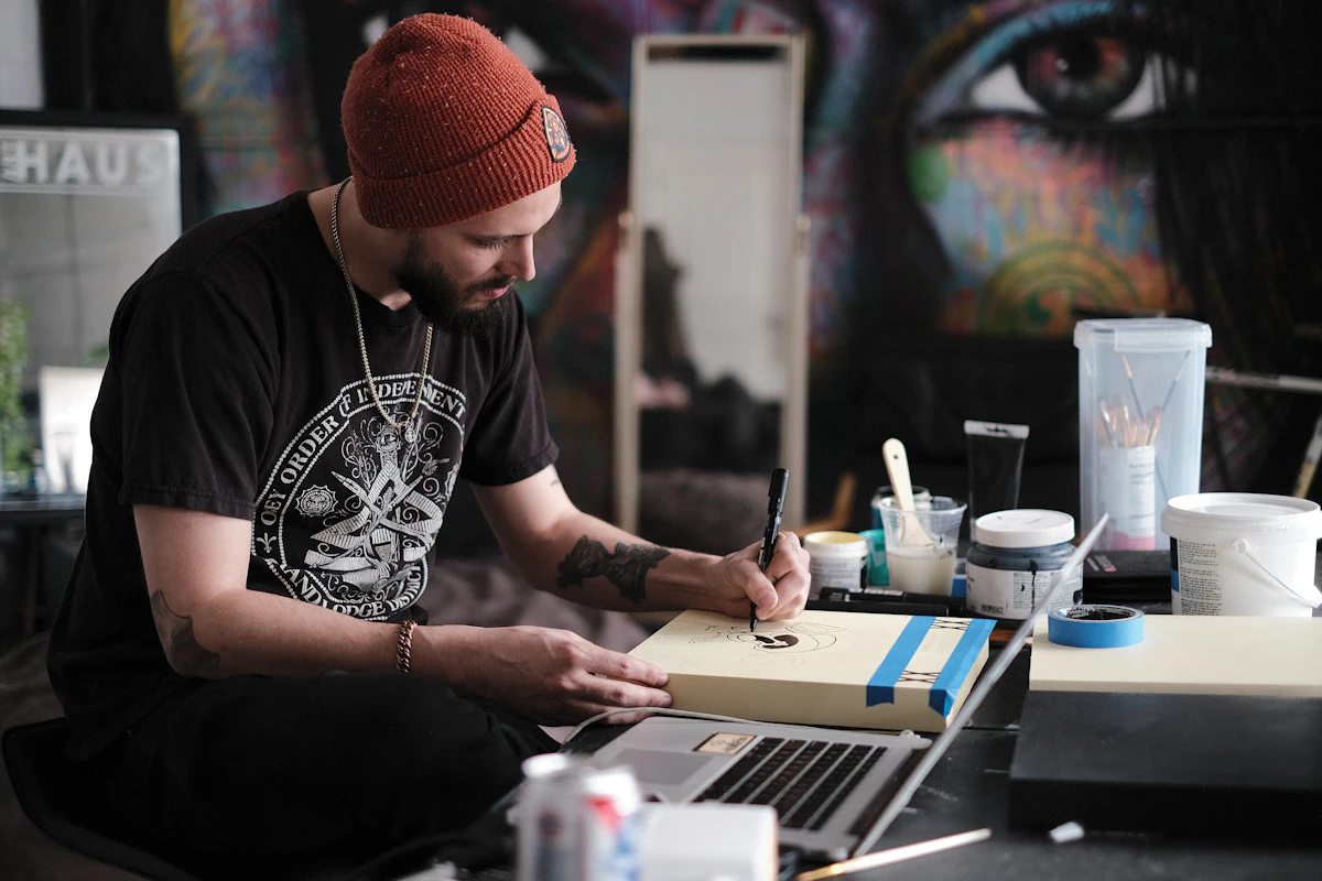 A tattoo artist in a beanie drawing a design at his desk, a mural of an eye behind him